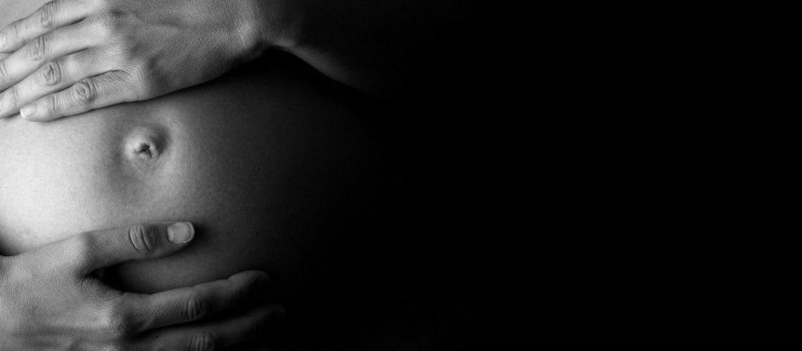 Artistic black and white close-up of an expectant mother holding her belly, concept of care, health and pregnancy, in low-key, with copy space on black.